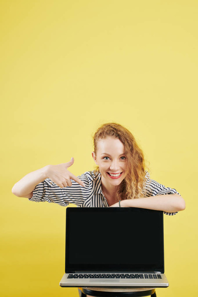 Pretty young smiling business lady pointing at black empty screen of opened laptop, isolated on yellow - Valokuva, kuva