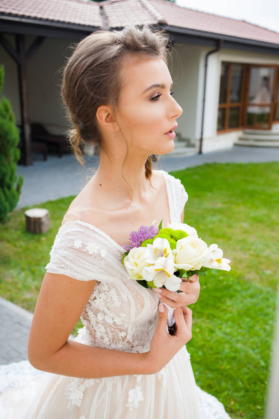 結婚式の前に白い家の近くを歩く光の長い白いドレスで結婚式の花束を持つ若いエレガントな女性. - 写真・画像