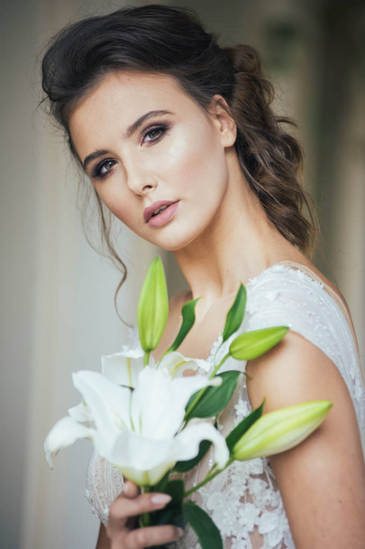 結婚式の前に白い家の近くを歩く光の長い白いドレスで結婚式の花束を持つ若いエレガントな女性. - 写真・画像
