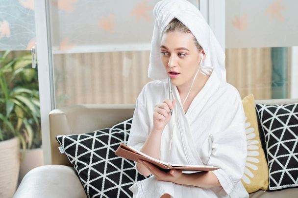 Young woman in bath robe listening to motivational audio book and writing down important thoughts and insights she had - Photo, Image