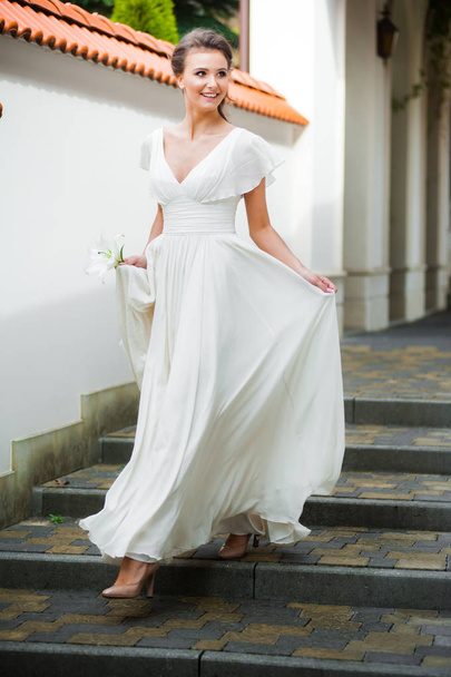 Νεαρή κομψή γυναίκα με γαμήλιο μπουκέτο σε ένα ελαφρύ μακρύ λευκό φόρεμα με τα πόδια κοντά στο λευκό σπίτι πριν από την τελετή του γάμου. - Φωτογραφία, εικόνα