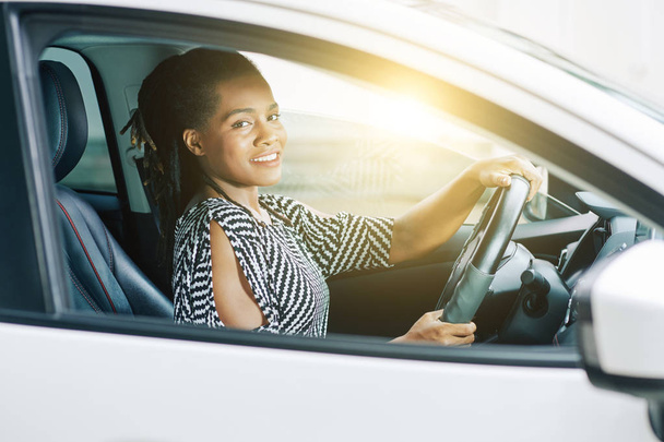 Muotokuva Afrikkalainen nuori nainen hymyilee kameralle istuessaan autossaan ja ajaessaan
 - Valokuva, kuva