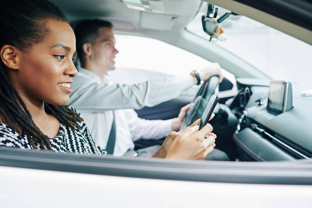 Afrykańska młoda kobieta czyta wiadomość przez telefon komórkowy i uśmiecha się podczas gdy młody mężczyzna prowadzi samochód - Zdjęcie, obraz