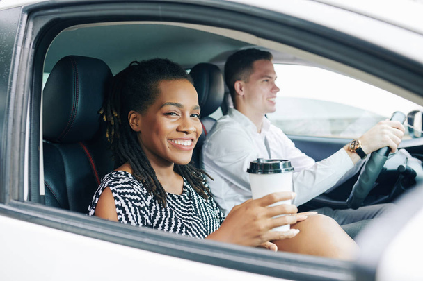 Mujer joven africana sonriendo a la cámara mientras bebe café durante su viaje en coche junto con un hombre joven
 - Foto, imagen