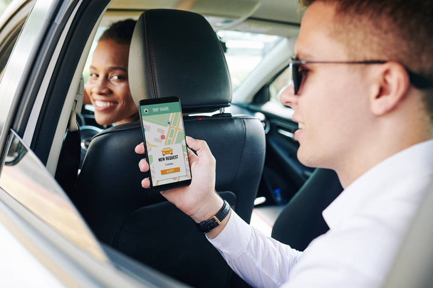 Hombre joven con gafas de sol sosteniendo el teléfono móvil con aplicación para compartir coche mientras está sentado en el asiento trasero con el conductor femenino sonriendo a la cámara
 - Foto, Imagen