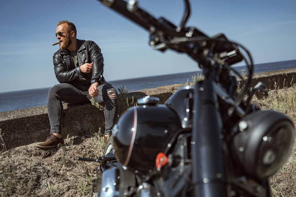 Cool bearded man in leather jacket sitting at the roadside by the ocean - Фото, зображення