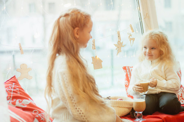 Two cute little sisters girls with blond curly hair  drink cocoa and eat gingerbread cookies by the window in anticipation of Christmas . Christmas mood - Φωτογραφία, εικόνα