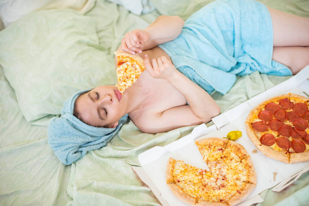 Cute girl with a towel on her head eats pizza in bed. Young woman eating pizza in bed. Life is a pleasure, body positive. Love for Italian food. Food habits, addiction to fast food. - Foto, Imagem