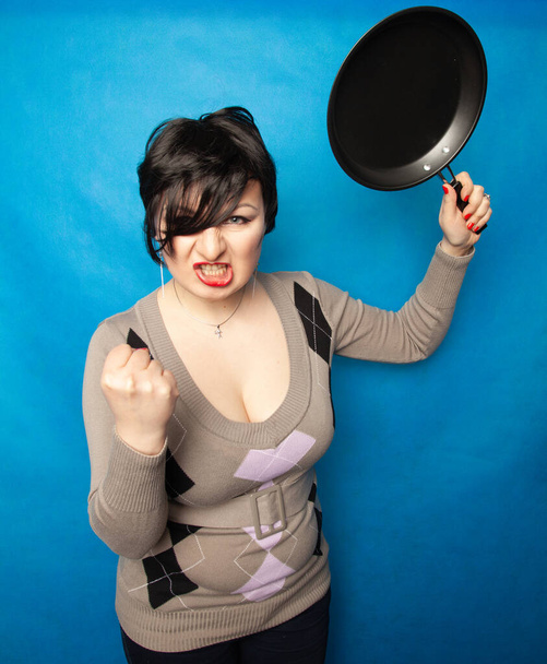 Woman beating with a frying pan on bluw studio background alone - Foto, afbeelding