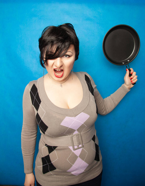 vrouw slaan met een koekenpan op bluw studio achtergrond alleen - Foto, afbeelding