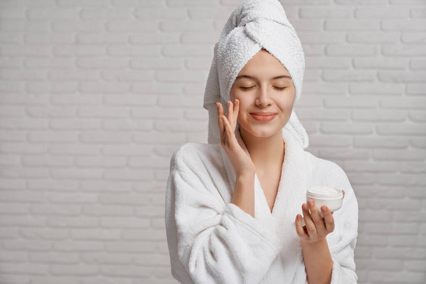 Young woman applying moisturizer on face with closed eyes. - Φωτογραφία, εικόνα