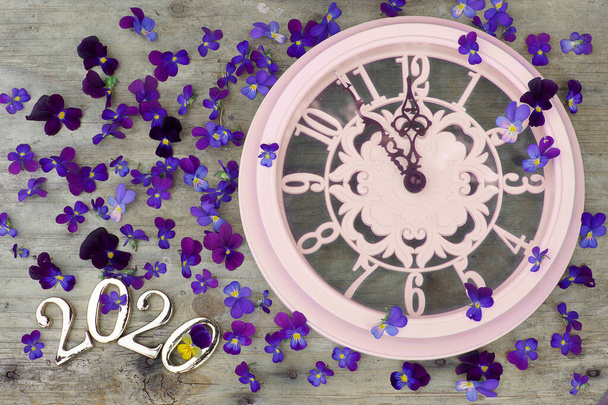 2020 numbers on a wooden background next to purple pansies flowers and a pink clock showing five minutes to midnight - Photo, Image