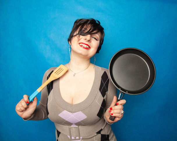 Front view portrait of a young happy caucasian female dressed in sweater, holding a frying pan in front of her, on blue studio background - Foto, afbeelding