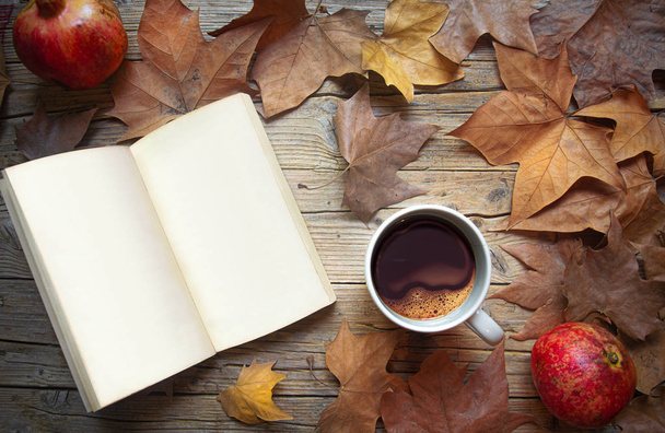 Παλιό ξύλινο τραπέζι με ανοιχτό βιβλίο και λευκές σελίδες, ξερά φθινοπωρινά φύλλα και φλιτζάνι καφέ - Φωτογραφία, εικόνα