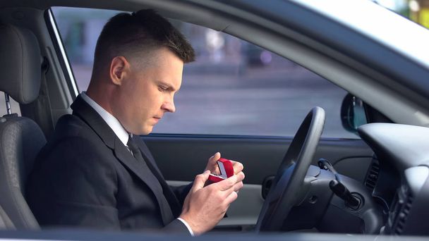 Красивый мужчина держит коробку с обручальным кольцом, сидит в машине, чувствуя себя неуверенно
 - Фото, изображение