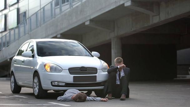 Απελπισμένος άντρας οδηγός κάθεται στο δρόμο κοντά στο αυτοκίνητο και αναίσθητος πεζός, κίνδυνος - Φωτογραφία, εικόνα