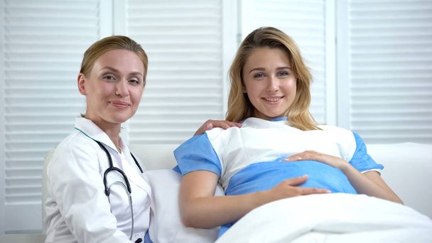 Gülümseyen hamile kadın ve jinekolog kameraya, doğum hastanesi ilanına bakıyor. - Fotoğraf, Görsel