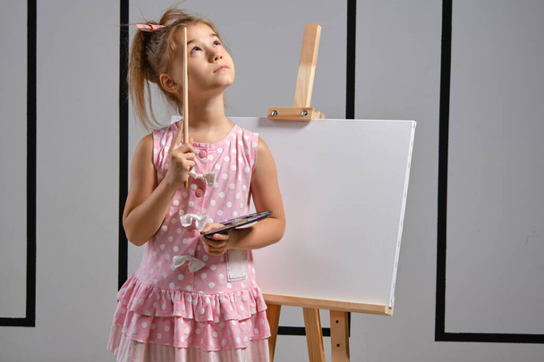 Petite fille artiste dans une robe rose est debout derrière le chevalet et la peinture au pinceau sur toile à l'atelier d'art avec des murs blancs. Plan rapproché moyen
. - Photo, image