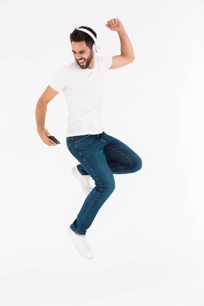 Full length image of excited smiling man dancing while listening music on cellphone and headphones - Foto, afbeelding