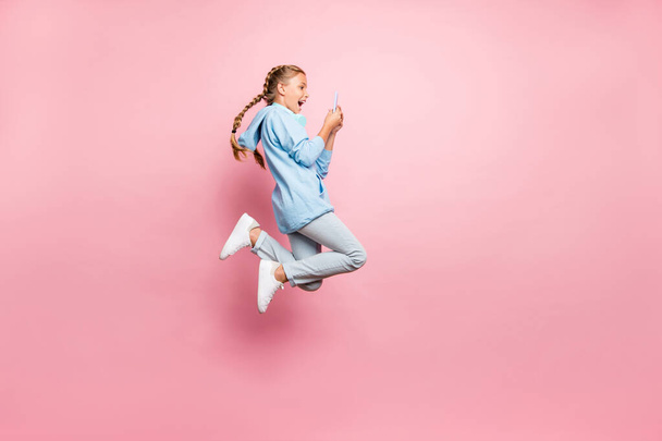 Ganzkörpergröße Foto von aufgeregt verrückt ekstatische Schulkind Browsing durch Telefon Springen tragen blaues Sweatshirt Pullover sportliche tragen Jeans Denim isoliert über rosa Pastellfarbe Hintergrund - Foto, Bild