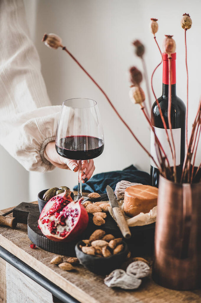 Red wine, board with cheese, fruit, almonds and olives, decorative flowers on kitchen counter and womans hand holding glass of wine, white wall at background. Wine tasting, wine bar concept - Foto, Bild