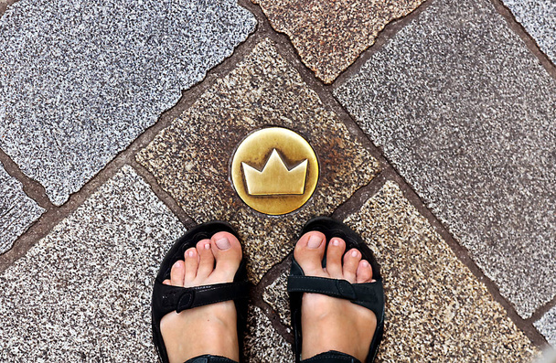 Horní pohled na unavené turistické nohy v sandálech a mosaznou korunu v dlážděných ulicích, která ukazuje umístění korunovační procházky, Bratislava, Slovensko. Staré město - Fotografie, Obrázek
