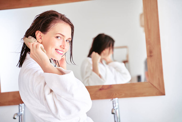 Photo of brunette woman combing her hair in front of bathroom mirror . - Foto, Bild