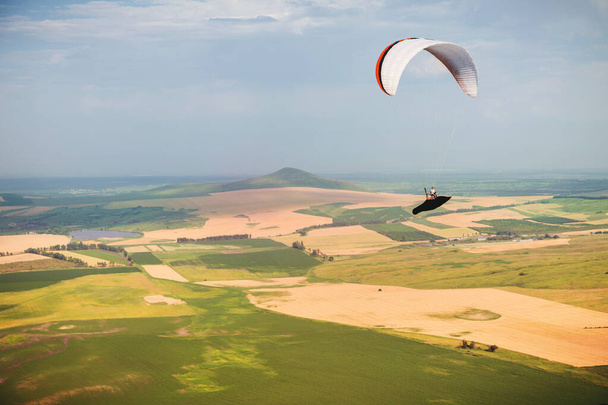 A paraglider flies in the sky in a cocoon suit on a paraglider over the Caucasian countryside with hills and mountains. Paragliding Sport Concept - 写真・画像