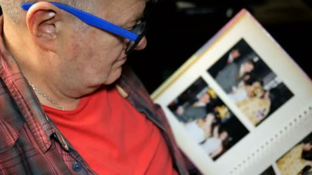 Starší muž kavkazského etnika, s modrými brýlemi, listuje albem s fotografiemi, sedí. Koncept vzpomínek a nostalgie ve středním věku. Zezadu. Detailní záběr. - Záběry, video