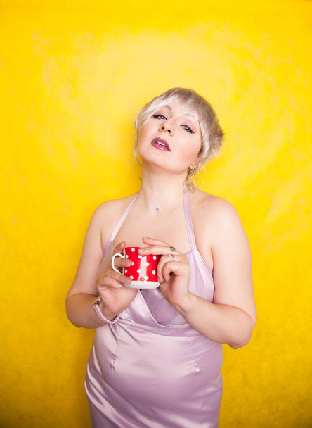bella curvy giovane donna bionda con i capelli corti gode di bere il tè da carino rosso a pois tazza di ceramica su sfondo giallo studio
 - Foto, immagini