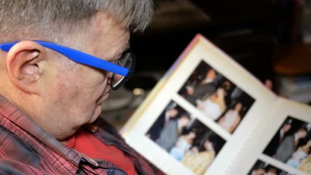 Senior man of Caucasian ethnicity, with blue glasses, leafs through album with photos, sitting. Concept of memories and nostalgia at middle-aged. From the back. Close-up. - Footage, Video