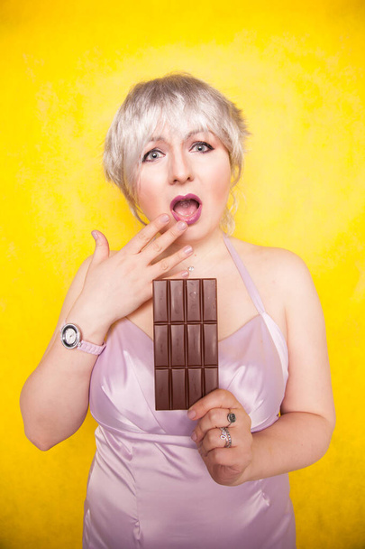 csinos szőke rövid haj nő a túlsúlyos gazdaság a kezében egy nagy csokoládé bár sárga fényes stúdió háttér - Fotó, kép