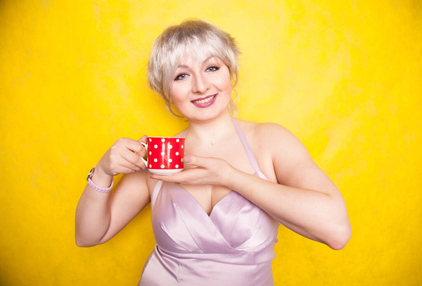 hübsche kurvige junge blonde Frau mit kurzen Haaren trinkt gerne Tee aus süßer roter Keramiktasse auf gelbem Studiohintergrund - Foto, Bild
