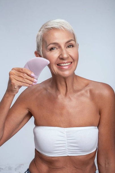Smiling mature lady using face cleaning brush - Fotó, kép