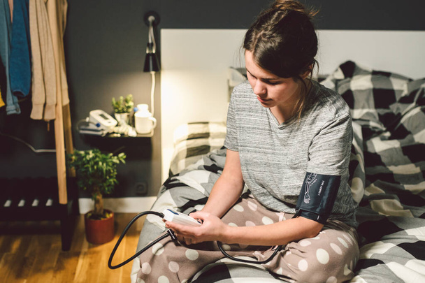 The topic of high blood pressure is hypertension disease. 22 year old young Caucasian woman uses an automatic tonometer to check pressure at home in the bedroom on the bed in home clothes in evening - Photo, Image