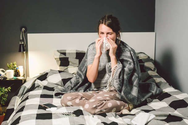 chora kobieta z katarem, siedząca w łóżku. Kobieta źle się czuje i kicha w sypialni. Kobieta trzymająca chusteczkę, objawy grypy. Koncepcja medyczna opieki zdrowotnej. Sezon zimna i grypy. kobieta z objawami alergii - Zdjęcie, obraz