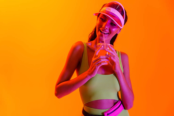 Emotional optimistic stylish girl in sport lifestyle clothes and transparent cap drinking juice cocktail isolated over orange background with neon lights. - Zdjęcie, obraz