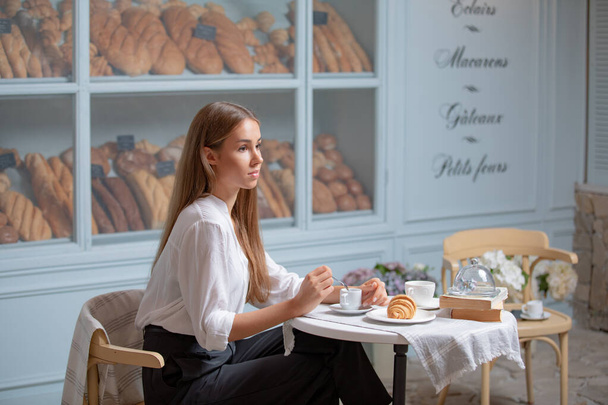 Žena si ráno dává snídani s croissantem a kávou. Velmi krásná dívka s dlouhými blond vlasy při obědě v pekárně - Fotografie, Obrázek