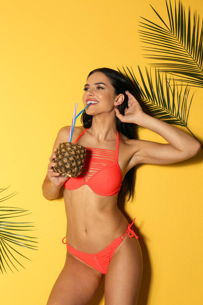 Beautiful young woman in stylish bikini with cocktail and tropical leaves on yellow background - Photo, Image