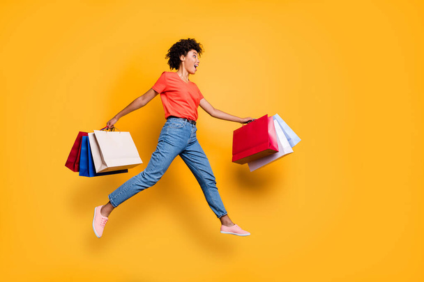 Full length profile side photo of amazed funky funny wavy brunette hair girl hold bags jump run to shopping center boutique hurry wear red t-shirt denim jeans isolated over yellow color background - Valokuva, kuva