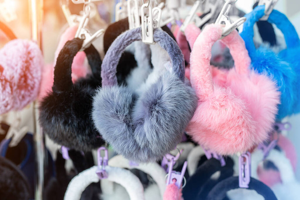 Много разноцветных ярких пушистых теплых зимних меховых наушников и перчаток висит на стойке на витрине магазина для продажи. Милые аксессуары для одежды холодного сезона
 - Фото, изображение