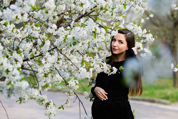 Chica embarazada en el jardín florecido. hermosa chica embarazada en un vestido negro está de pie en un jardín florecido
 - Foto, imagen