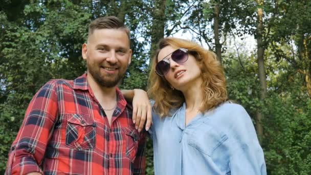 Lovely couple of bearded hipster boyfriend in red shirt and blond girlfriend in blue casual jeans blouse looking at the camera and putting on his dark sunglasses on green trees background - Footage, Video