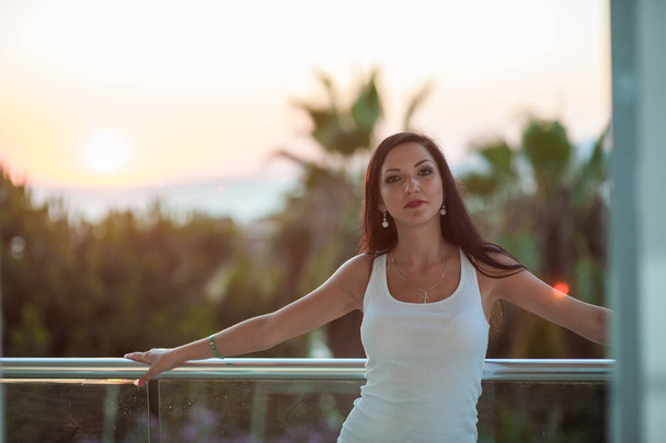 The girl on the background of the sea sunset. beautiful brunette at sunset sea vacation enjoying the warm sunshine. beautiful model outside with the sun in the background in a white t-shirt - Fotografie, Obrázek