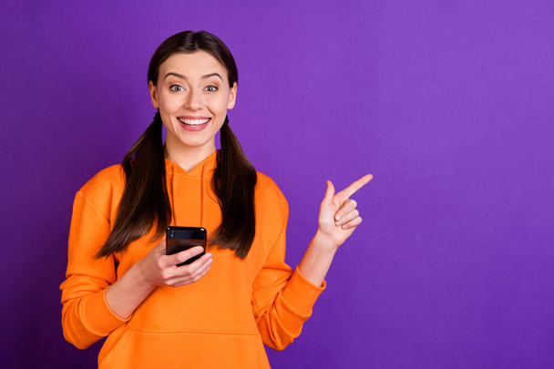 Portrait of positive cheerful youth girl blogger hold her cellphone point indexf inger on copyspace repost news wear orange trendy stylish sportswear isolated over violet purple color background - Foto, afbeelding