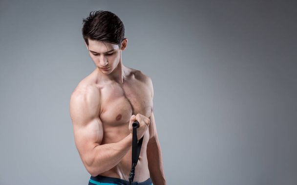 Muscular fitness jóvenes hombres antiguos músculos perfectos seis paquetes de abdominales y pecho desnudo. El modelo Bodybuilder entrena con un elástico de estiramiento sobre un fondo oscuro en el estudio. Gimnasio de entrenamiento
 - Foto, Imagen