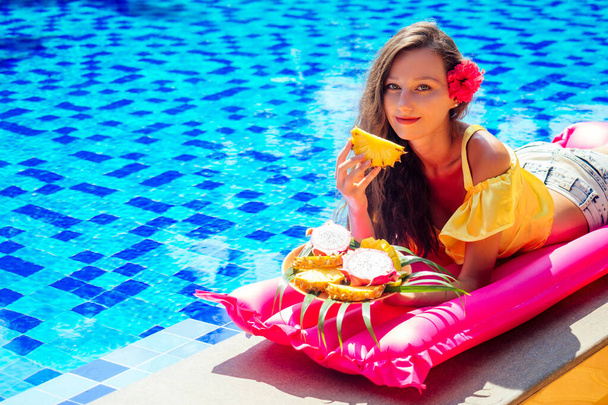menina bonita que encontra-se em um colchão cor-de-rosa inflável que come a manga das frutas tropicais, a fruta do dragão, o mamão e o abacaxi em um prato perto da piscina.
 - Foto, Imagem