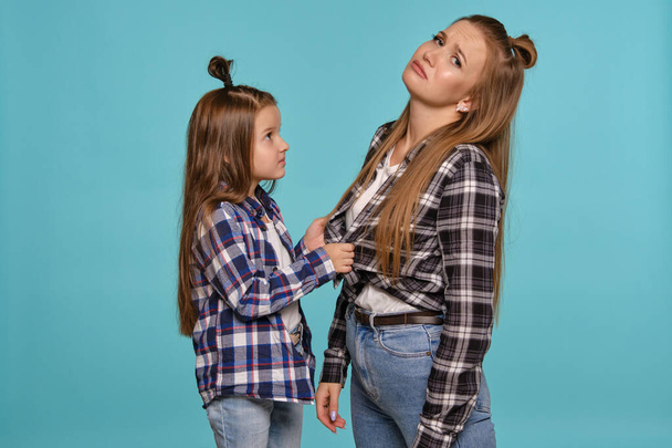 Mom and daughter with a funny hairstyles, dressed checkered shirts and blue denim jeans are posing against a blue studio background. Close-up shot. - Photo, Image