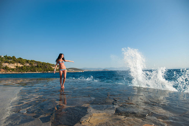 A girl in a swimsuit, splashing from the waves, Brunette girl rejoices splashing from the waves. Young happy woman jumping with raised arms, waving a blue bikini swimsuit in a blue swimsuit and a spla - Photo, Image