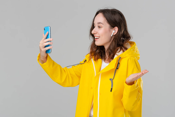 Όμορφη μελαχρινή γυναίκα σε ένα βίντεο με κίτρινο αδιάβροχο κουβεντιάζοντας στο smartphone απομονωμένο σε γκρι φόντο. Έννοια της επικοινωνίας. - Φωτογραφία, εικόνα